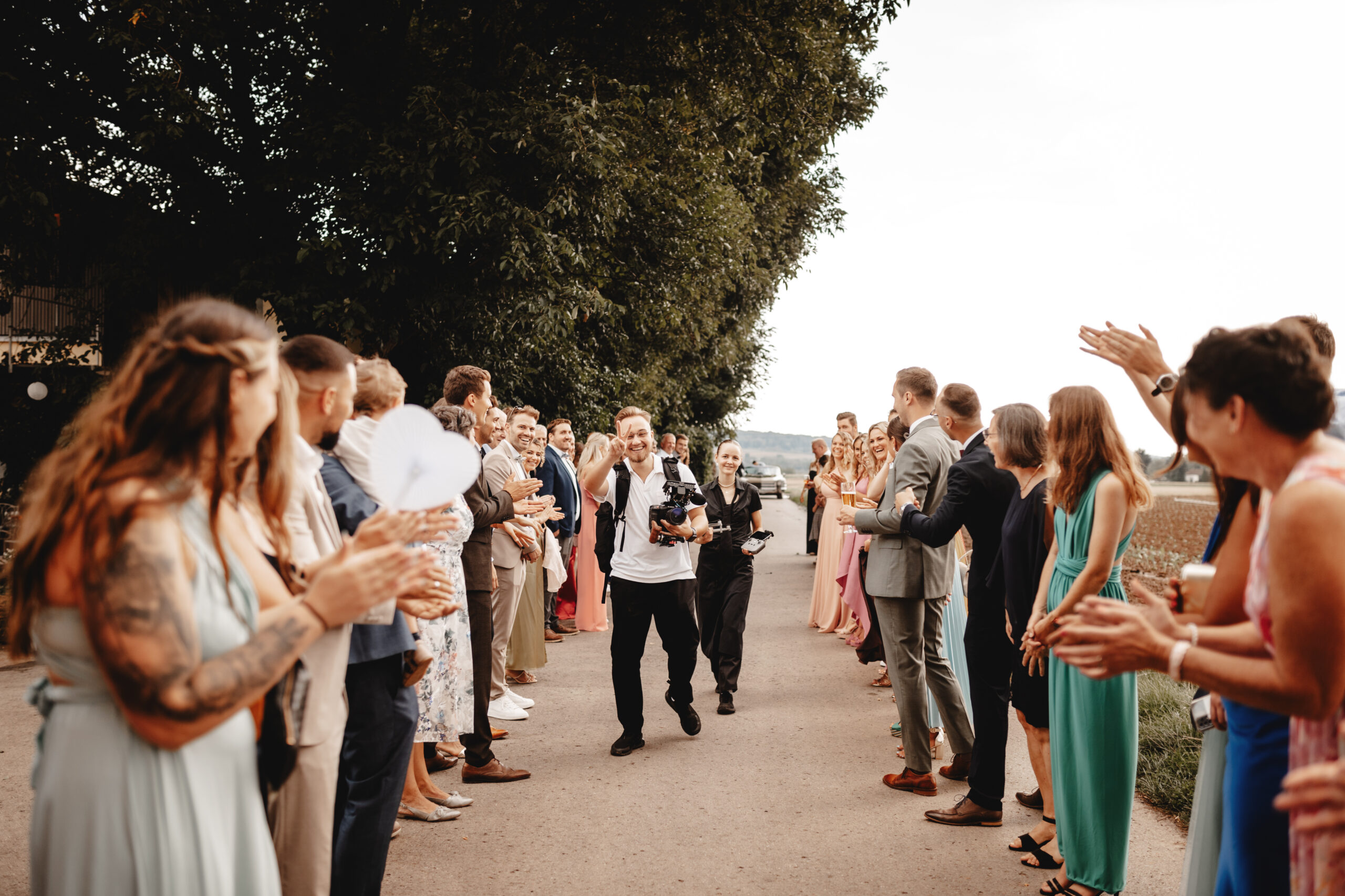 Videograf beim Hochzeitsvideo Stuttgart läuft lachend mit Kamera durch das Spalier der Gäste, die ihn aus Spaß anfeuern, bevor das Brautpaar folgt.