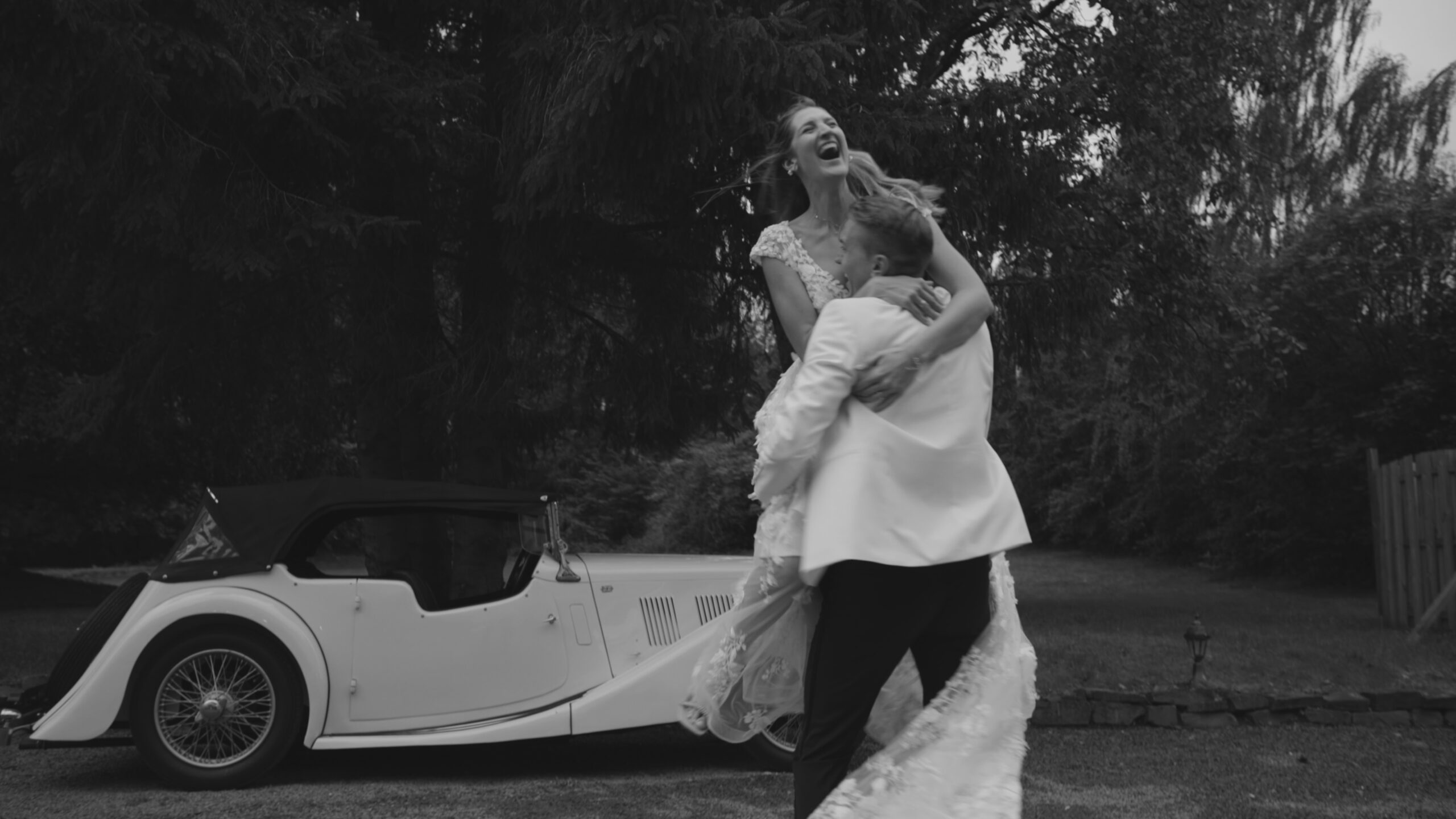 Romantischer Moment bei einem Hochzeitsvideo Stuttgart: Der Bräutigam hebt die Braut hoch und dreht sich mit ihr wie in einer Filmszene, im Hintergrund ein weißes Oldtimer-Auto, Schwarz-Weiß-Aufnahme.